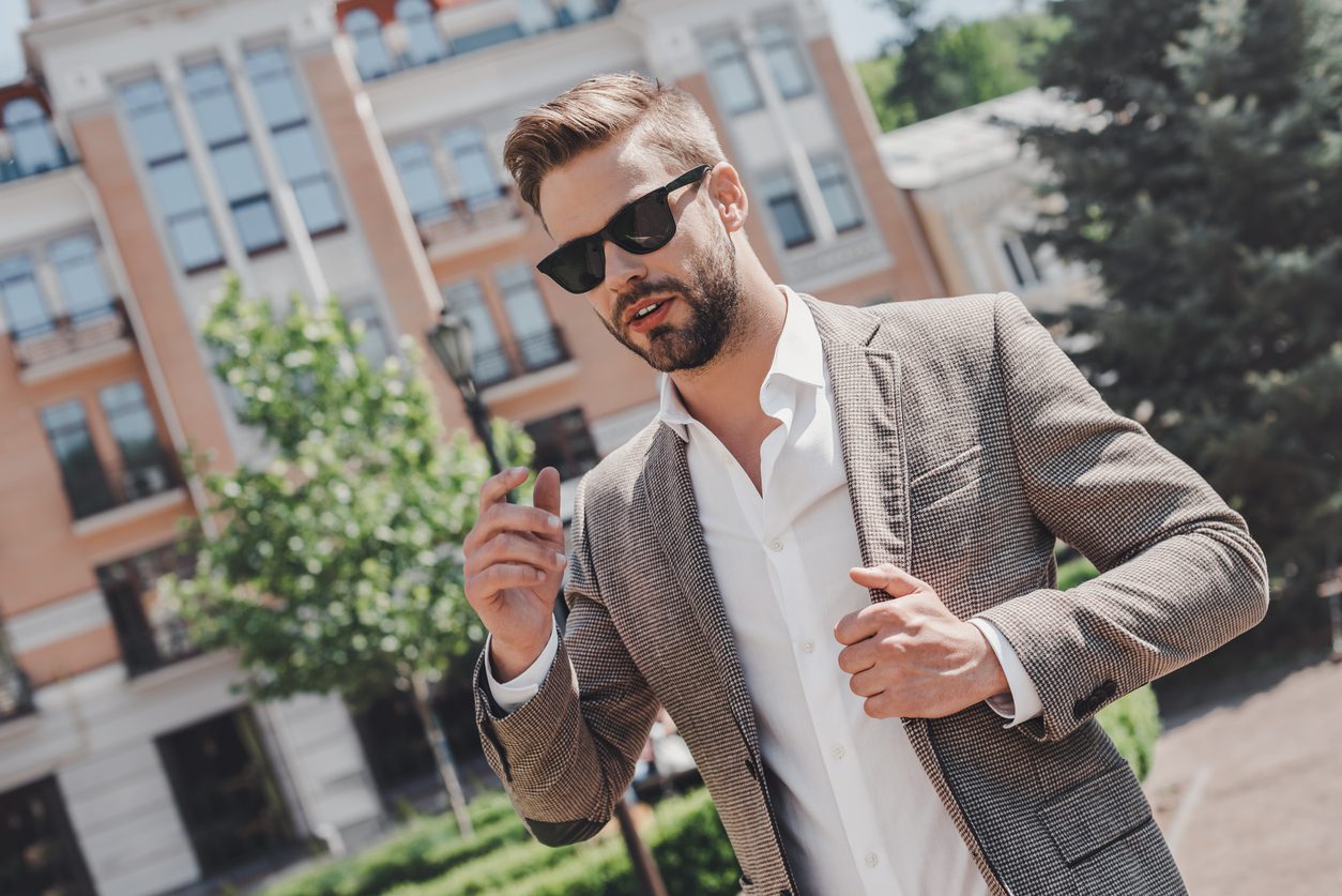 Man wearing a brown jacket, white shirt, and sunglasses from his summer wardrobe outdoors while standing on the city street.